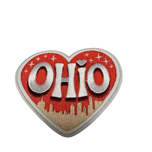 Pode incluir: Um emblema bordado em forma de coração com fundo vermelho, estrelas brancas e a palavra OHIO em letras grandes prateadas. Uma silhueta dourada do horizonte urbano está bordada na parte inferior, encimada por um pequeno coração prateado.