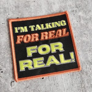 May include: A black embroidered patch with orange stitching. The patch says "I'm talking for real for real!" in a yellow and gray font.