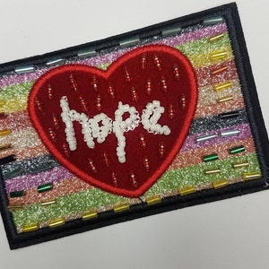 May include: A red heart-shaped patch with the word "Hope" spelled out in white beads on a multi-colored glittery background.