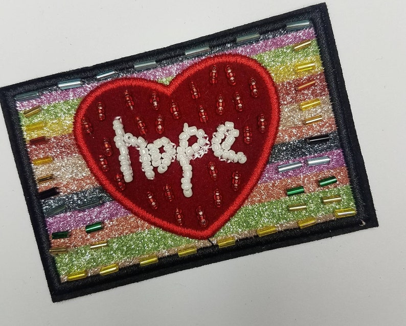 May include: A red heart-shaped patch with the word "Hope" spelled out in white beads on a multi-coloured glittery background.