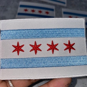 Può includere: Una toppa ricamata bianca con la bandiera di Chicago. La bandiera presenta quattro stelle rosse su una striscia blu chiaro su sfondo bianco.