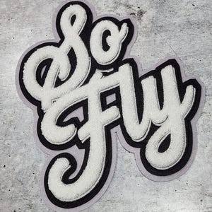 Może przedstawiać: Biała łatka z filcu z czarnym obramowaniem, z napisem "So Fly" w stylizowanym, ozdobnym piśmie.