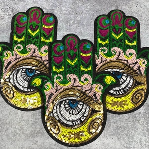 以下が含まれることがあります： 緑、金、ピンクの布地のパッチが3つあり、手と目のデザインが施されています。パッチはスパンコールで飾られています。