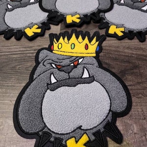 May include: Embroidered patches featuring a bulldog wearing a gold crown. The patches are primarily gray with black and yellow accents. The bulldog has a stern expression and the letter "K" is featured.