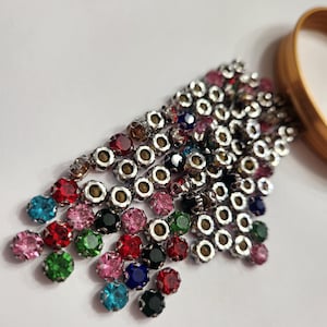 Peut inclure: Une collection de strass colorés dans diverses nuances de rouge, rose, vert, bleu et noir. Les strass sont sertis dans des montures en métal argenté. Un anneau de couleur or est visible en arrière-plan.