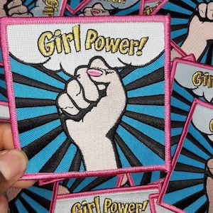 Könnte beinhalten: Ein Bügelbild mit einer erhobenen Faust und dem Text "Girl Power!" in goldenen Buchstaben auf blauem und rosa Hintergrund.