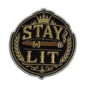 Puede incluir: Un parche bordado circular con fondo negro y borde dorado. Presenta una corona dorada en la parte superior, el texto STAY LIT en dorado y un cigarro marrón con una banda dorada en el centro. Hojas de laurel doradas enmarcan los lados del diseño.