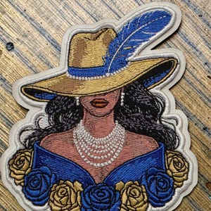 Könnte beinhalten: Gestickter Aufnäher mit einer Frau, die einen breitkrempigen Hut mit einer blauen Feder, eine Perlenkette und ein blaues Kleid trägt. Das Design umfasst goldene und blaue Rosen mit einem weißen Rand.