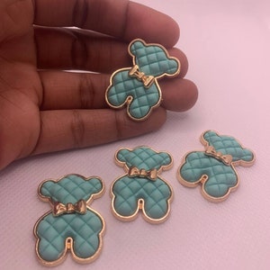 Op de afbeelding: Vier tealblauwe teddybeer-vormige harsversieringen met gouden rand en een gouden strik. De teddyberen hebben een gestikte textuur.