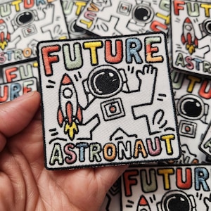 May include: White embroidered patch with the words "FUTURE ASTRONAUT" in colorful letters. The patch features a cartoon astronaut with a rocket ship. Other patches are visible in the background.