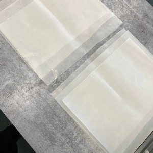 May include: A stack of four clear, translucent sheets of parchment paper. The sheets are arranged in a staggered pattern on a grey concrete surface.