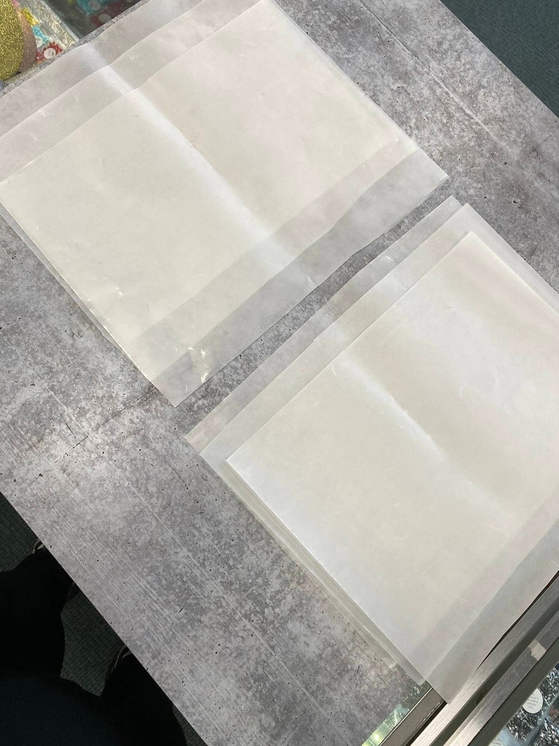 May include: A stack of four clear, translucent sheets of parchment paper. The sheets are arranged in a staggered pattern on a grey concrete surface.