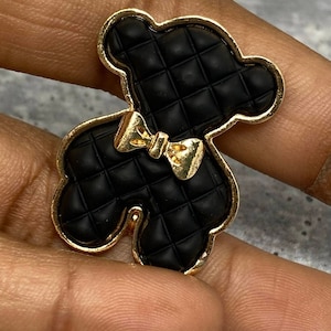 Op de afbeelding: Een zwarte teddybeer-vormige bedel met een gewatteerde textuur en een gouden strik. De bedel heeft een gouden omtrek, waarschijnlijk voor sieraden of accessoires.