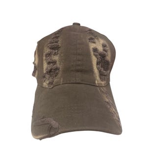 May include: A dark brown baseball cap featuring a distressed, worn-in aesthetic with frayed edges and intentional rips across the front panels and the curved brim. The fabric displays a faded, uneven color texture throughout the entire surface.