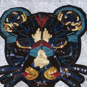 Pode incluir: Um patch bordado preto em forma de rosto de urso. O patch é adornado com lantejoulas douradas, azuis e vermelhas. As orelhas e as características faciais do urso são delineadas com lantejoulas azuis e douradas.