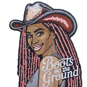 May include: Embroidered patch featuring a smiling woman with pink braids and a cowboy hat. The hat is brown and silver. The text "Boots on the Ground" is on the woman's shoulder. The patch is on a white background.