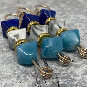 May include: Three gold safety pins with blue and white marble-patterned glass beads. The beads are square-shaped and have gold accents.