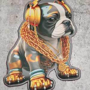 Puede incluir: Un bulldog francés blanco y negro con auriculares dorados y un collar de cadena de oro. El perro está sentado sobre un fondo gris.