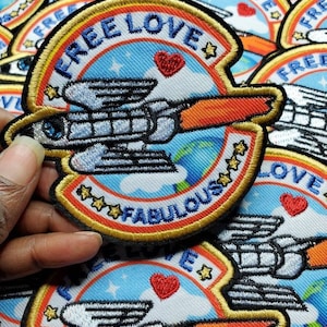 Könnte beinhalten: Gestickter Aufnäher mit den Worten "FREE LOVE" und "FABULOUS" in einem kreisförmigen Design. Der Aufnäher zeigt eine Rakete, Sterne und ein Herz. Der Hintergrund ist blau mit Wolken und einem Globus.