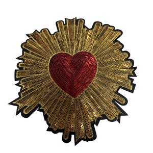 Puede incluir: Un parche decorativo que presenta un corazón rojo central cubierto de lentejuelas, rodeado por un patrón radiante de lentejuelas doradas. Todo el diseño está delineado con un borde negro, creando un contraste definido con los colores metálicos.