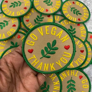 Op de afbeelding: Een verzameling ronde, bruine stoffen patches met groene stiksels. De patches hebben een groen bladerenontwerp en de tekst "Go Vegan Thank You" in gele letters. Er zijn rode harten op de patches.