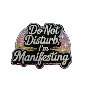 May include: An embroidered patch featuring the text "Do Not Disturb, I'm Manifesting" in white script and block lettering. The background is a black arch shape with a pastel rainbow gradient, small white star accents, and a pink border outline.