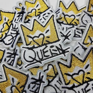 May include: Embroidered patches featuring a gold crown design with a heart and the word "QUEEN" in black graffiti-style lettering. The patches have a white background and a gray border. The image shows a pile of these patches.