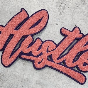 Puede incluir: Un parche de fieltro rosa y morado con la palabra "Hustle" escrita en una fuente estilizada.