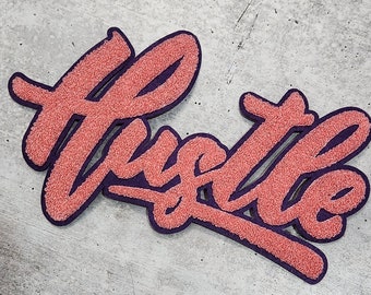 May include: A pink and purple felt patch with the word "Hustle" written in a stylised font.