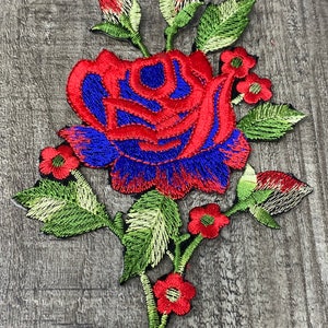 May include: A red and blue embroidered rose with green leaves and stems. The rose has a single bloom with smaller red flowers surrounding it.