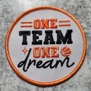 以下が含まれることがあります： オレンジと黒で「ONE TEAM ONE dream」の文字が刺繍された円形のパッチ。パッチはオレンジ色の縁取りとバスケットボールのグラフィックが特徴です。背景は白です。