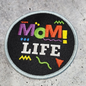 May include: A black and white embroidered patch with the text "MOM! LIFE" in a colorful 90s style font. The patch has a light blue border.