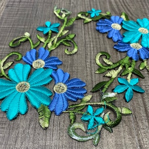 May include: A floral applique with blue and green flowers and leaves. The applique is made of fabric and is designed to be sewn onto a garment or other fabric.