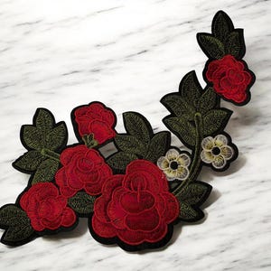 Könnte beinhalten: Gestickte Blumenapplikation mit roten Rosen, grünen Blättern und kleinen weißen und gelben Blüten. Die Applikation hat einen schwarzen Rand und eignet sich zum Verzieren von Kleidung oder Bastelprojekten.