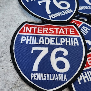 May include: A blue and red Interstate 76 Philadelphia Pennsylvania highway sign patch. The sign is a white oval with the text "Interstate" in red, "Philadelphia" in white, and "76" in white. The bottom of the sign says "Pennsylvania" in white.