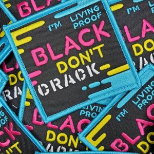 Może przedstawiać: Zbliżenie na kilka czarnych łat z tkaniny z tekstem "I'm Living Proof Black Don't Crack" w różowych, żółtych i białych literach. Łaty mają niebieską obwódkę i są ułożone jedna na drugiej.