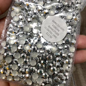 May include: A clear plastic bag filled with silver Korean rhinestuds. The rhinestuds are 6mm in size and have a faceted, reflective surface. A white circular label with black text indicates the product details.