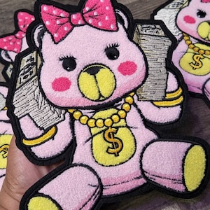 Op de afbeelding: Roze pluche teddybeer met een gouden ketting met een dollartekenhanger en een roze strik met witte stippen. De beer houdt een stapel geld in zijn linkerpoot en draagt een gouden armband om zijn rechterpoot.