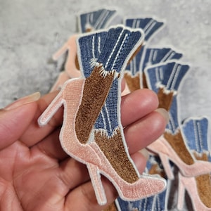 May include: Embroidered patches featuring a high-heeled shoe design. The patches depict a pair of legs in blue denim jeans and brown boots, with a pink high heel. The patches are made of fabric and thread.