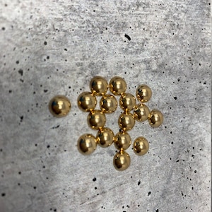 Könnte beinhalten: Eine Gruppe von 20 goldfarbenen runden Metallperlen, verstreut auf einer grauen, strukturierten Oberfläche.