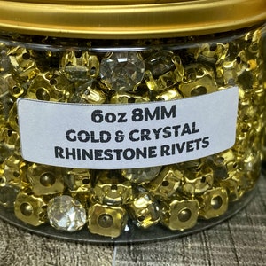 Peut inclure: Un bocal transparent rempli de 170 g de rivets en strass dorés et transparents de 8 mm. Le bocal a un couvercle doré et une étiquette blanche sur laquelle est inscrit "6oz 8MM GOLD & CRYSTAL RHINESTONE RIVETS".