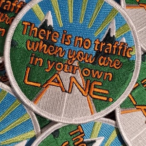 Pode incluir: Um remendo branco redondo com um design bordado em azul, verde e laranja. O remendo apresenta uma estrada com montanhas ao fundo e o texto "There is no traffic when you are in your own LANE."