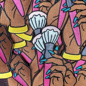 Op de afbeelding: Een set geborduurde patches met handen die make-upkwasten vasthouden. De kwasten hebben roze en blauwe handvatten en witte haren. De handen zijn bruin met gele armbanden en blauwe vingernagels.