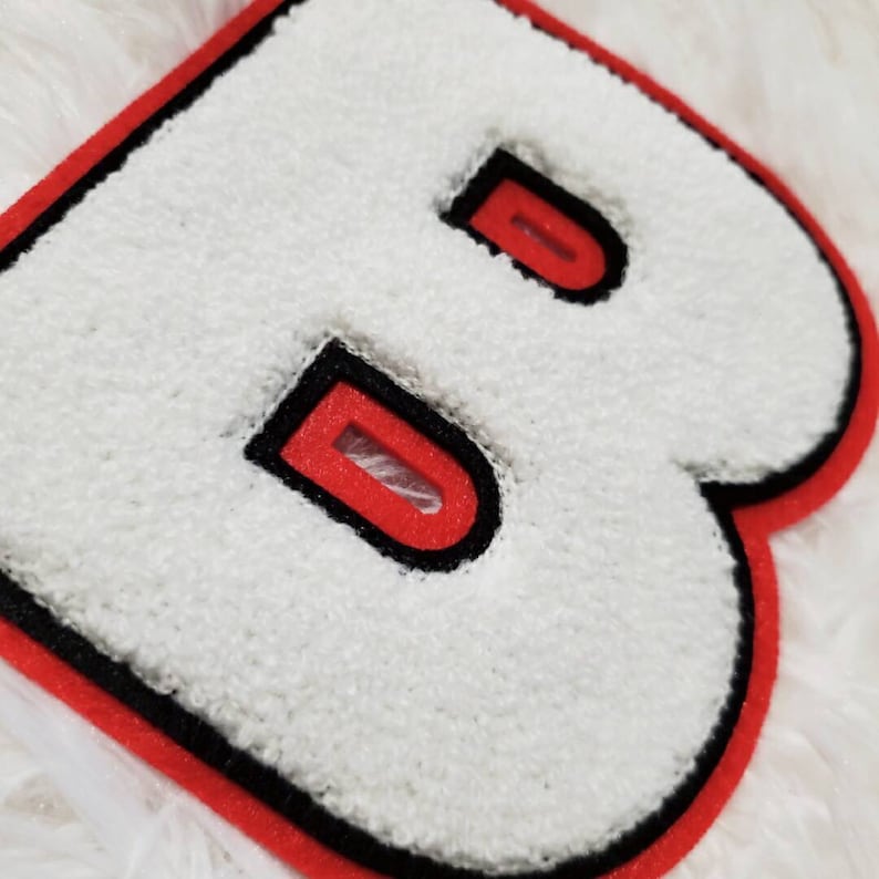 Puede incluir: Letra B de fieltro blanco con contorno rojo y negro. La letra es gruesa y tiene un efecto 3D.