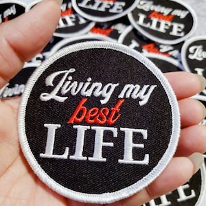 May include: A black and white embroidered patch with a white border. The patch is round and features the text "Living my best LIFE" in white with the word "best" in red.