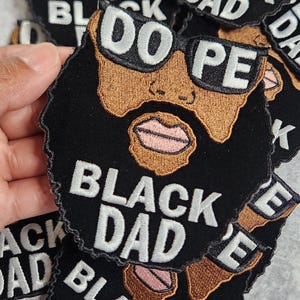Puede incluir: Parche bordado negro y marrón con una ilustración de dibujos animados de un hombre negro con gafas de sol y barba. El texto "Dope" está por encima de la cabeza del hombre y el texto "Black Dad" está por debajo de la cabeza del hombre.