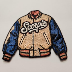 Puede incluir: Un parche bordado de una chaqueta de béisbol beige y azul con la palabra "Scorpio" en letras blancas en la parte delantera.