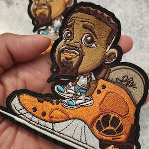 May include: Embroidered patch featuring a cartoon character wearing a white shirt and blue shoes, sitting on a large orange shoe with a black and white sole. The character has brown skin, brown hair, and a brown beard. The patch is on a black background.