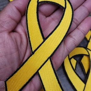 Puede incluir: Parche de cinta amarilla con borde negro. La cinta está doblada en forma de lazo. El parche está hecho de tela y tiene una superficie texturizada.