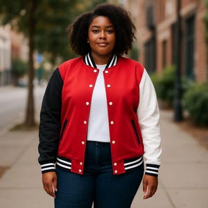 May include: A red, black, and white varsity jacket with gold buttons. The jacket has black sleeves on the left and white on the right. The jacket is worn over a white t-shirt and blue jeans. The jacket has black and white striped cuffs and hem.